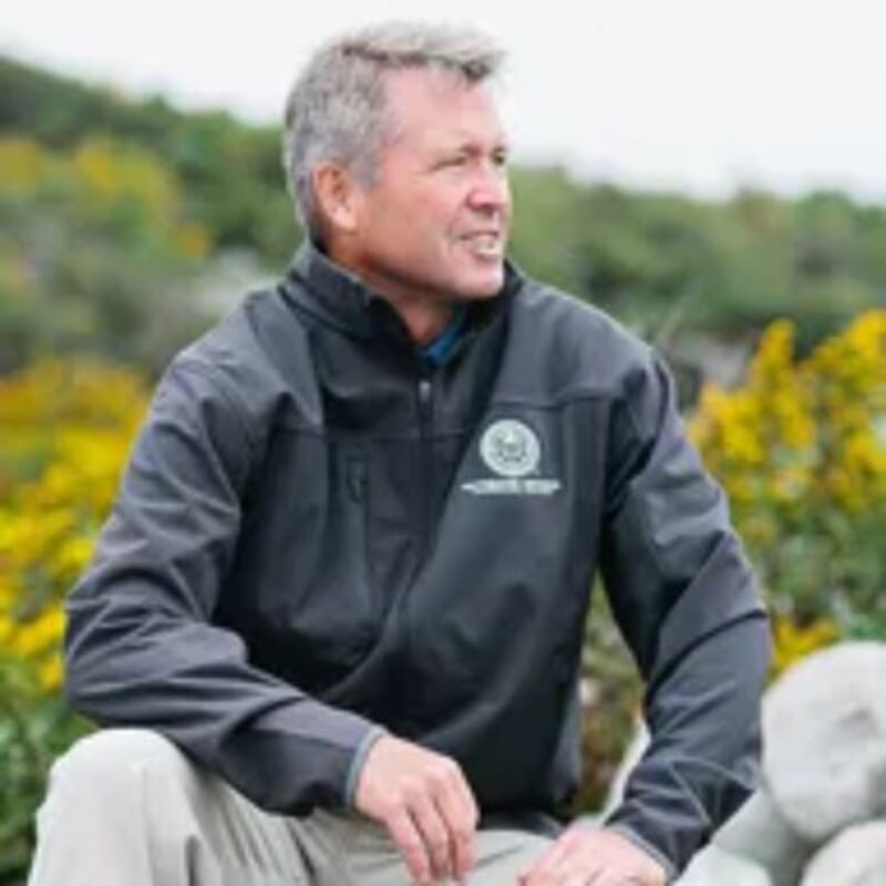 The image shows a middle-aged man with short gray hair, wearing a dark jacket and light-colored pants. He is sitting outside with a blurred background of greenery and yellow flowers. The man is looking off to the side with a slight smile. The jacket has a logo on the chest.